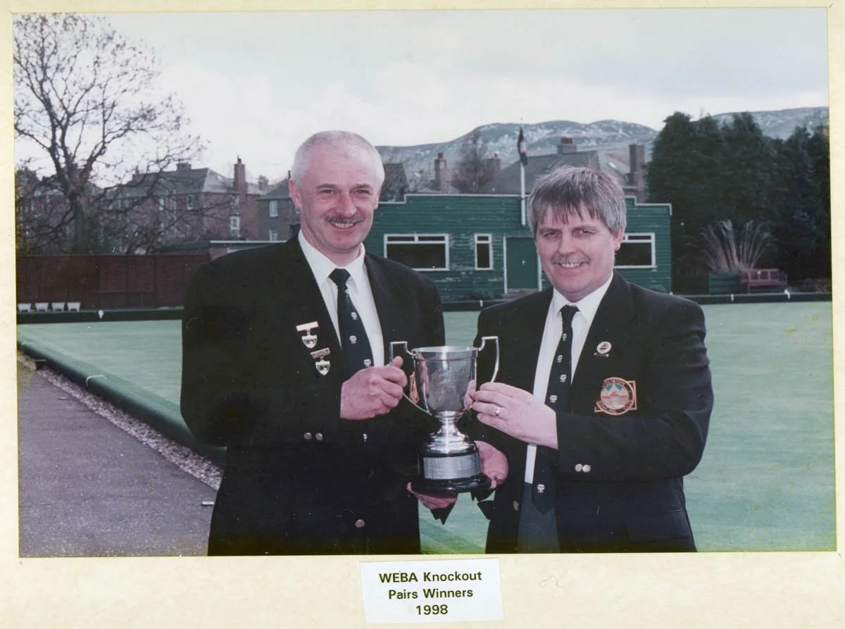 Two men in suits holding a trophy, standing outdoors on a lawn with a building and hillside in the background. The photo is labeled 'WEBA Knockout Pairs Winners 1998'.