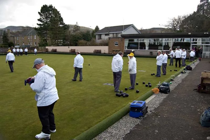 People playing lawn bowls on a grass field outside a building with large windows, surrounded by trees and hills in the background.