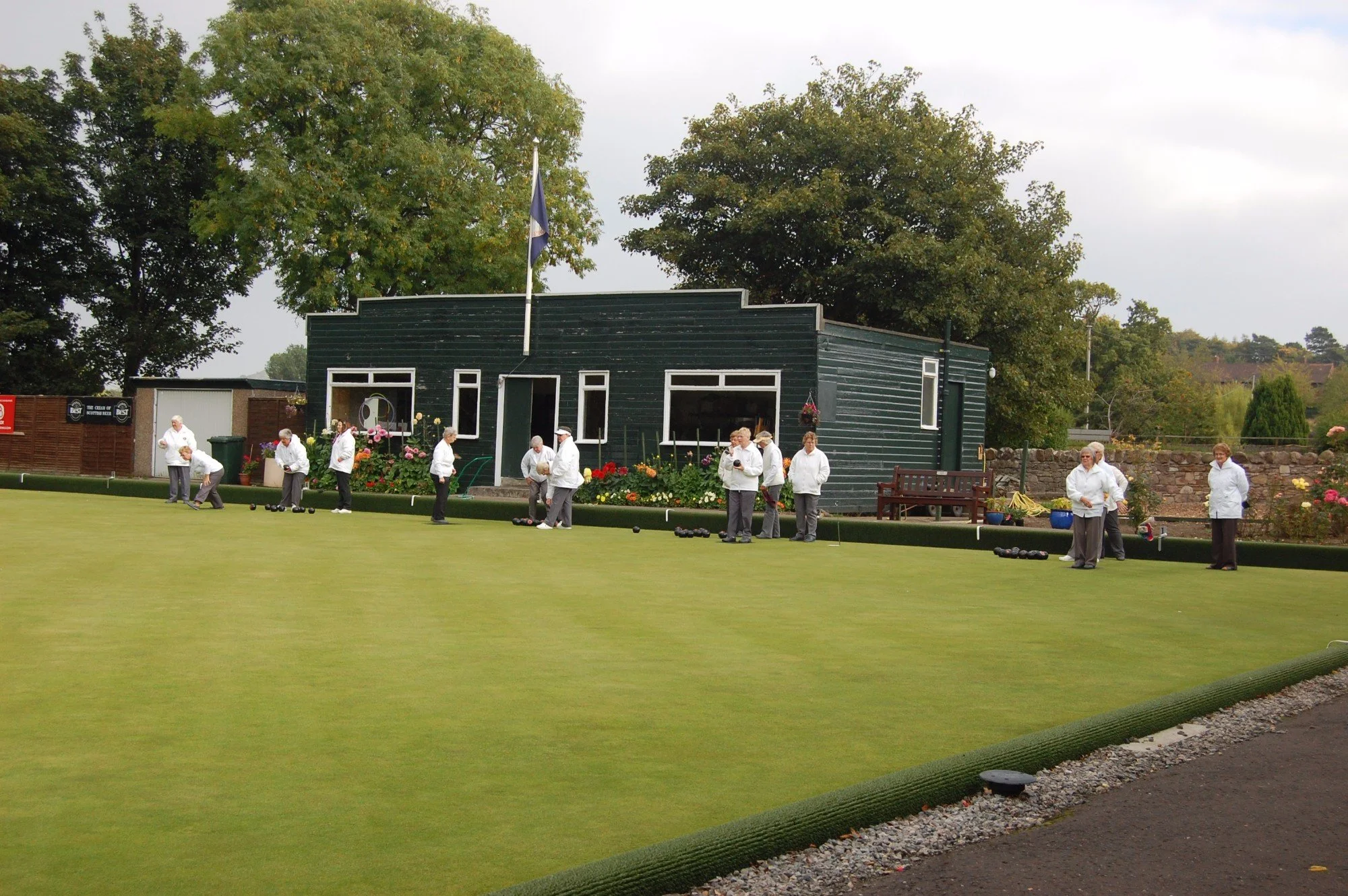 A group of elderly people dressed in white jackets playing bowls on a green lawn at a park or club, with trees and a dark wooden building in the background.