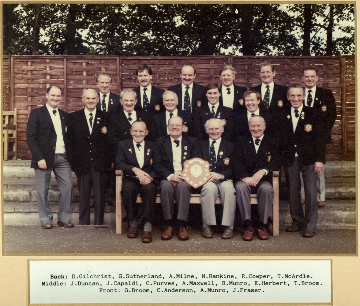 A group photo of 18 men dressed in police or military uniforms, posing outdoors in front of a wooden fence, with five seated men in the front row holding a shield. The photo is printed with a caption listing their names and positions.