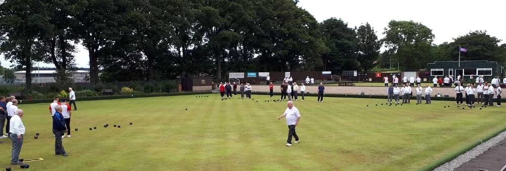 People playing and watching a game of lawn bowls on a well-maintained outdoor lawn, with trees and social areas in the background.