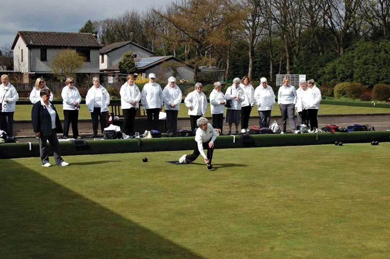A group of women playing lawn bowls outdoors on a cloudy day, with some spectators watching behind a low barrier.