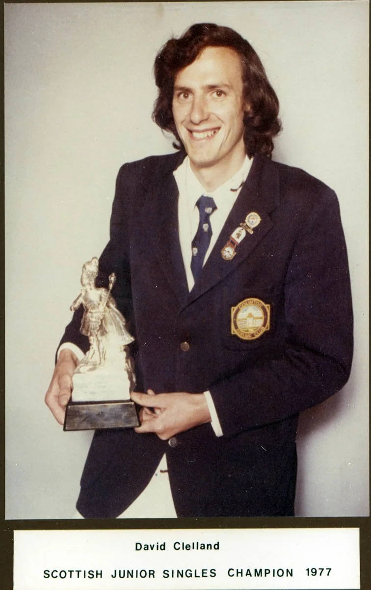 A man in a suit holding a trophy, smiling at the camera. The trophy depicts a figure in a sports pose. There are pins on his lapel and an emblem on his blazer. The text below reads, "David Clelland, Scottish Junior Singles Champion 1977."