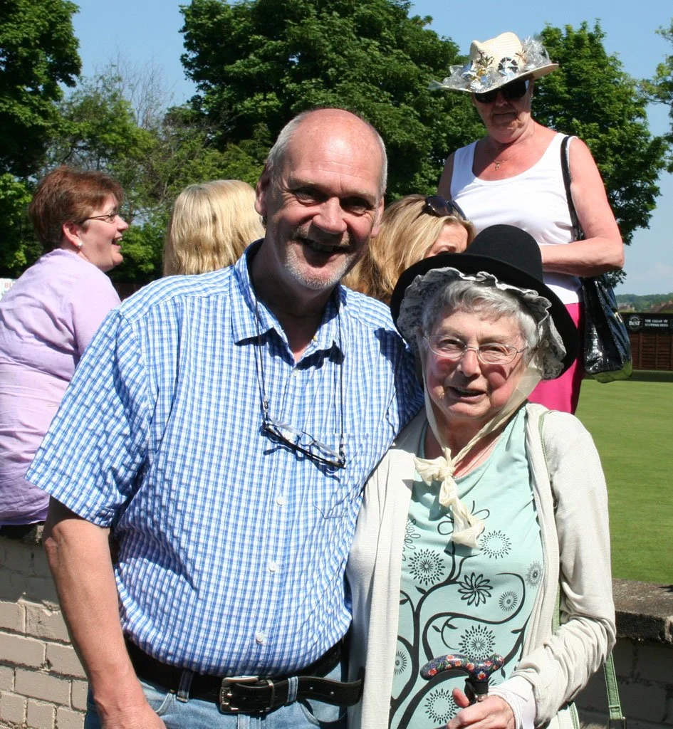 Group of joyful elderly people outdoors, smiling, with one woman wearing a large hat and glasses, and a man with glasses hanging from his shirt, standing on a grassy area under trees on a sunny day.