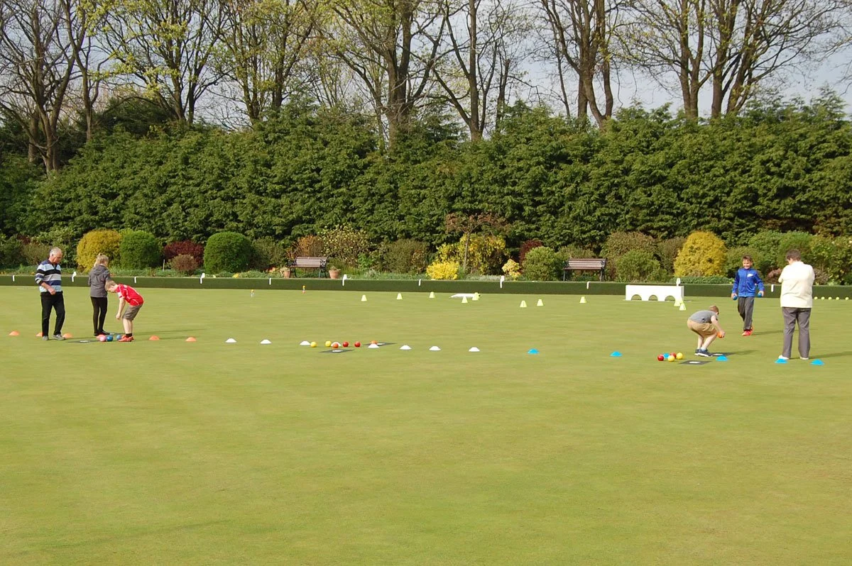 People playing lawn bowls on a neatly trimmed grass field with green bushes and trees in the background.