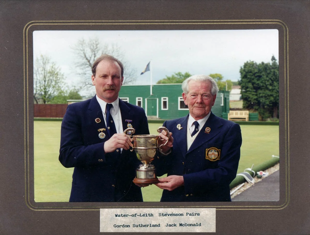 Two men in formal uniforms are holding a large trophy together outdoors on a golf course, with a green building, trees, and a flag in the background.