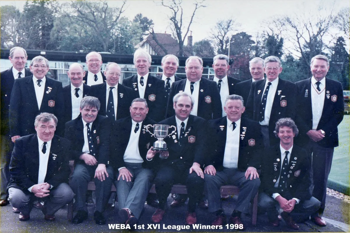Group of men in black suits with white shirts and ties, some have badges, posing outdoors with one man holding a trophy. The caption reads 'WEBA 1st XVI League Winners 1998'.