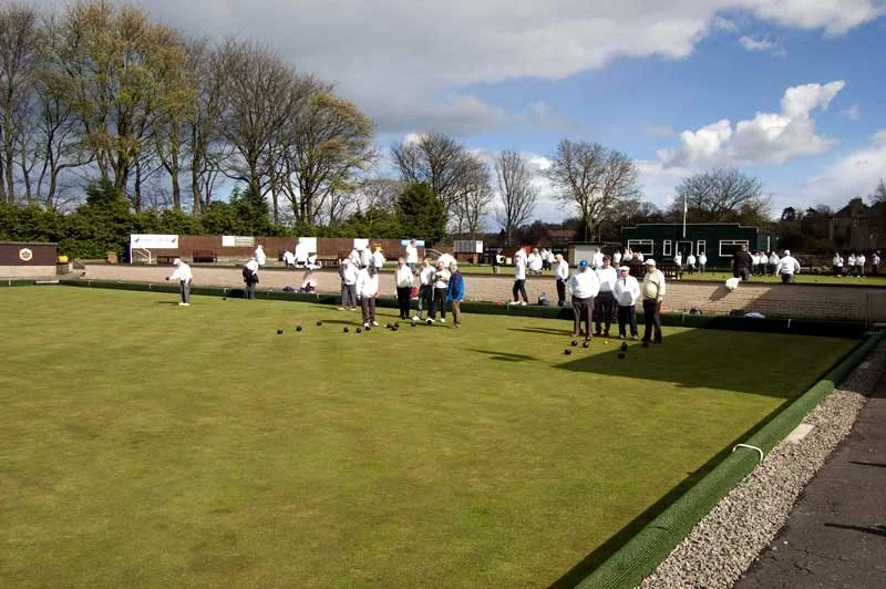 People playing lawn bowling on a grassy outdoor field with trees and a blue sky in the background.