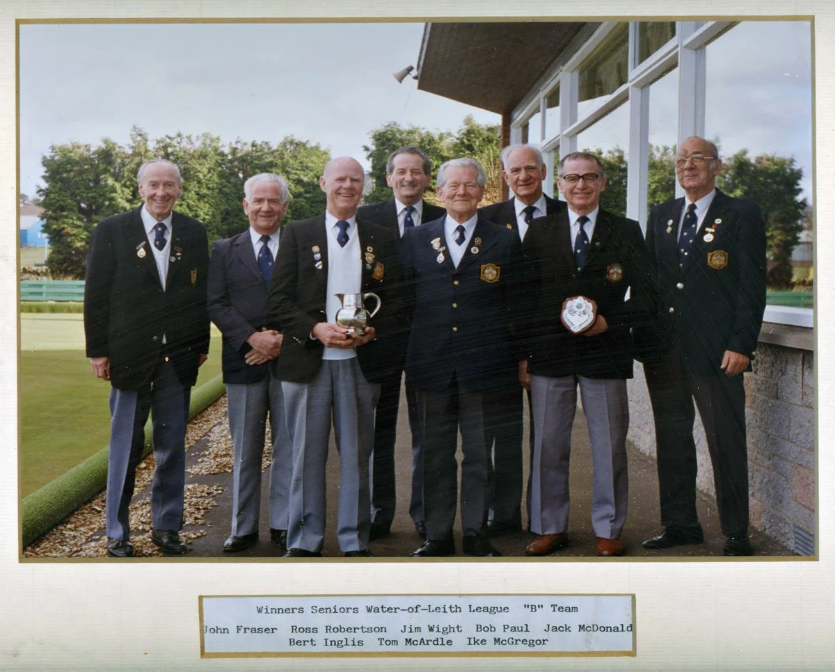 Group of nine older men in suits with badges, standing outside next to a building with large glass windows, holding trophies and medals in a celebratory pose, with trees in the background.
