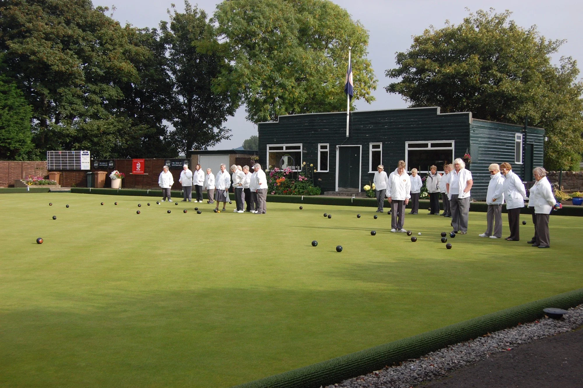 A group of women dressed in white jackets and dark pants are playing lawn bowls on a neatly mowed green field, with a building and tall trees in the background.