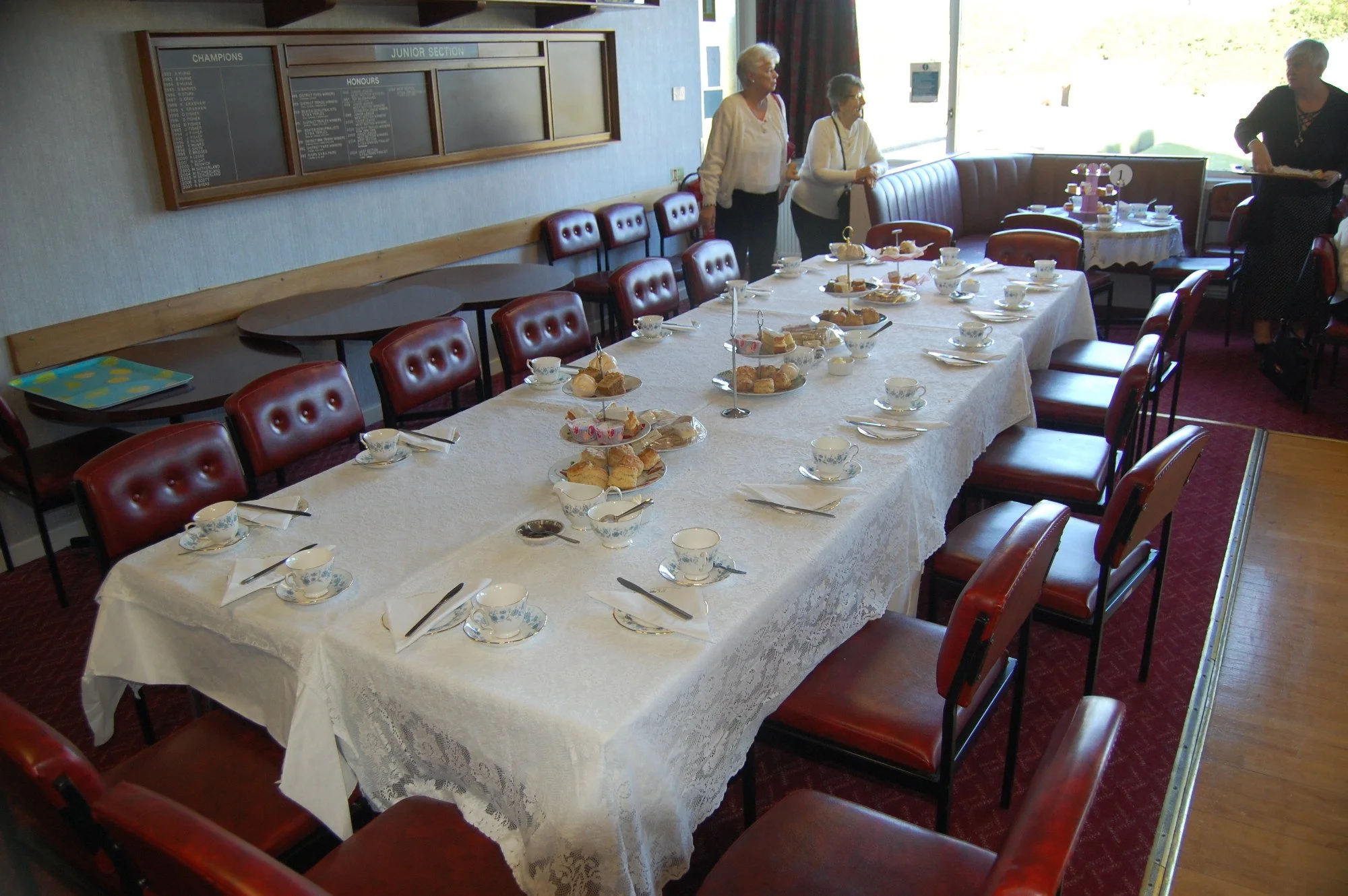 A dining room set up for an event with a long table covered in a white lace tablecloth, decorated with tea cups, plates, and tiered trays of assorted pastries and cakes. Several women are standing near a window, and the room features red cushioned ch