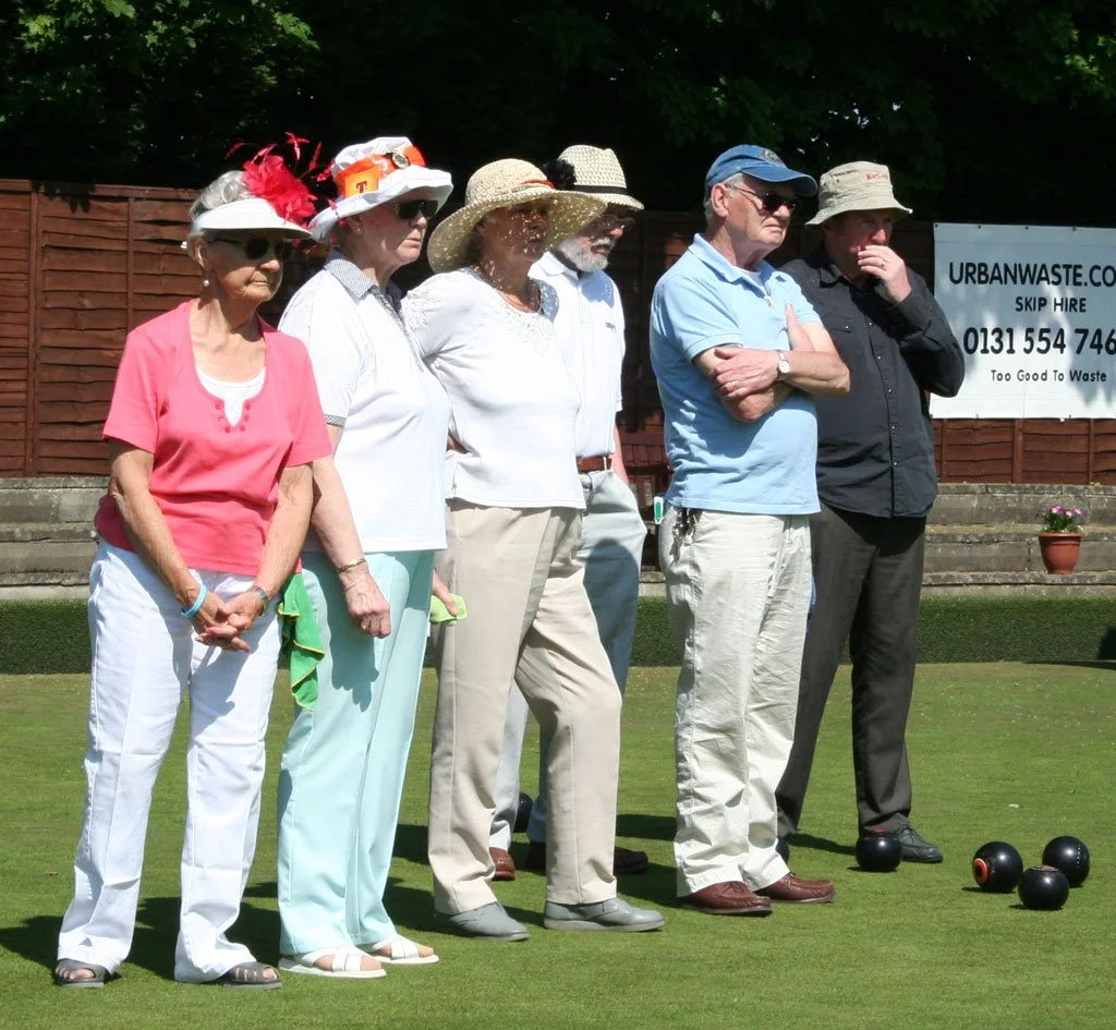 Group of six elderly people standing on a lawn, participating in a bocce game. They are wearing casual summer clothing and hats, with some wearing sunglasses. Four black bocce balls are on the ground in front of them. A brown wooden fence and a banne