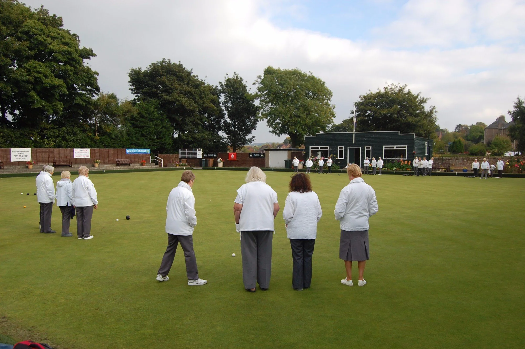 People dressed in white jackets are playing lawn bowls on a green outdoor field, with a small dark building, trees, and sky in the background.