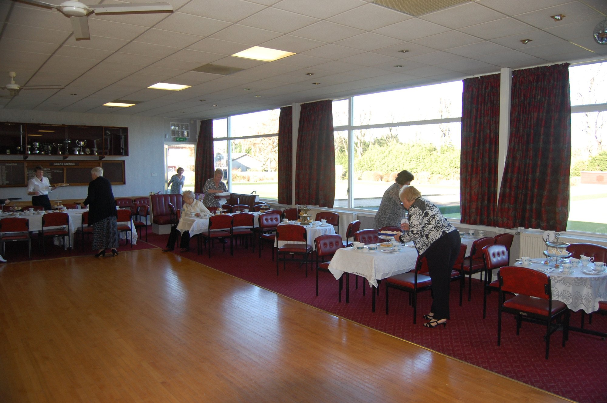 People preparing and serving food in a decorated dining hall with large windows and red curtains during the daytime.