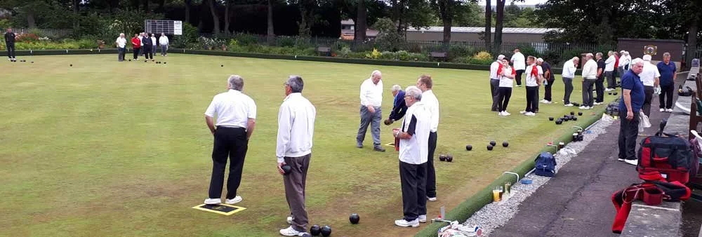 People playing and watching lawn bowls on a green surrounded by trees and benches.