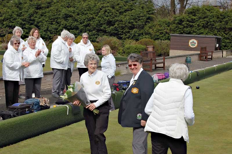 A group of mostly elderly women gathered on a lawn at a park, some holding flowers and clapping, with many wearing white jackets. One woman in the foreground is smiling and holding flowers, while another woman has her back to the camera. There is a s