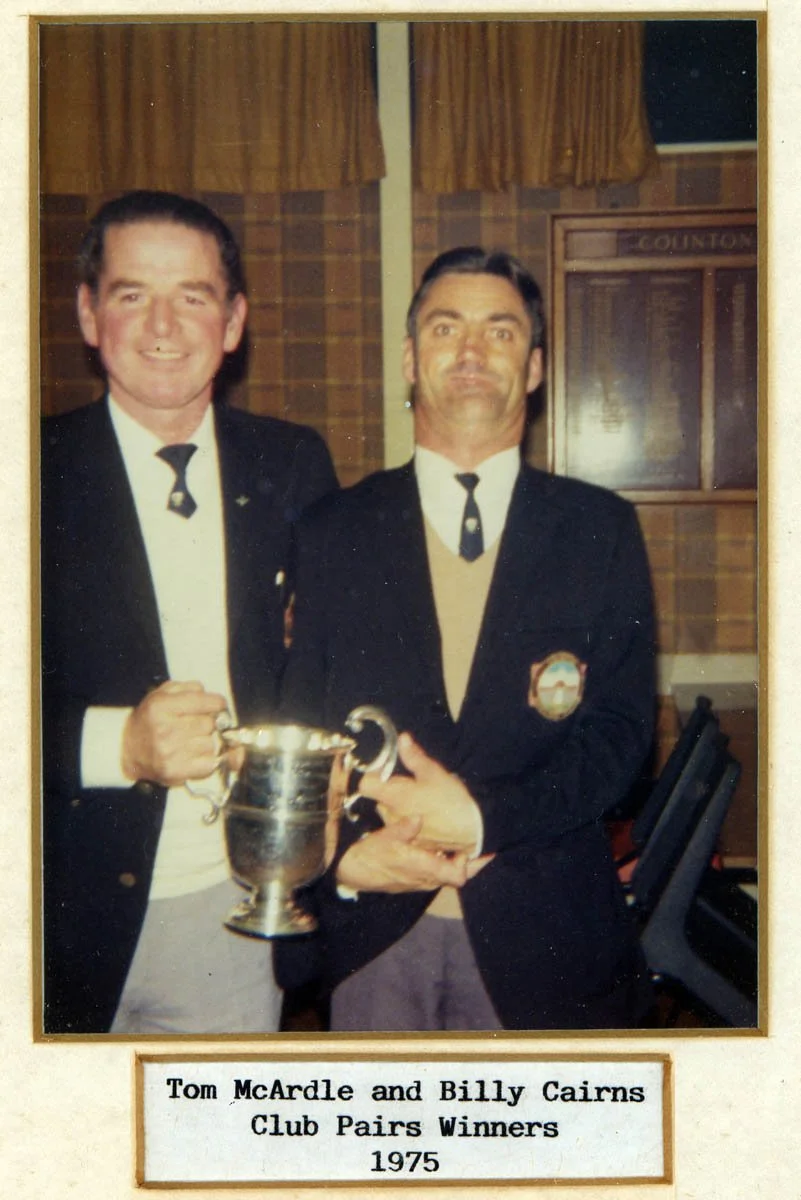 Two men in formal attire holding a trophy, standing in a room with wood-paneled walls and a plaque. The label below indicates they are Tom McArdle and Billy Cairns, Club Pairs Winners in 1975.