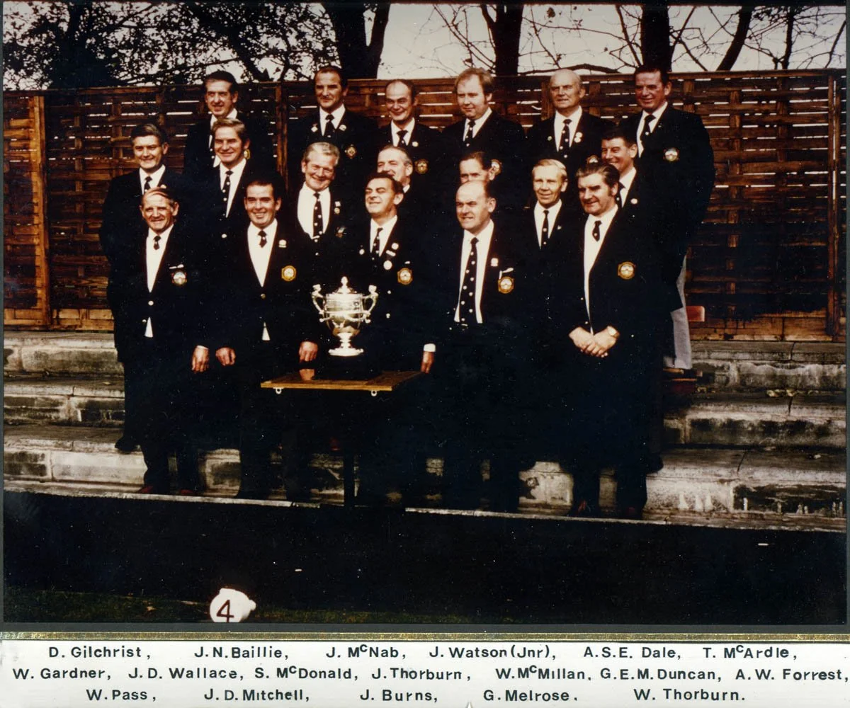 A black-and-white photograph of a group of men dressed in formal suits with badges, posing outdoors behind a wooden fence with leafless trees behind them. They are standing on a set of concrete steps, and a table with a large trophy is in front of th