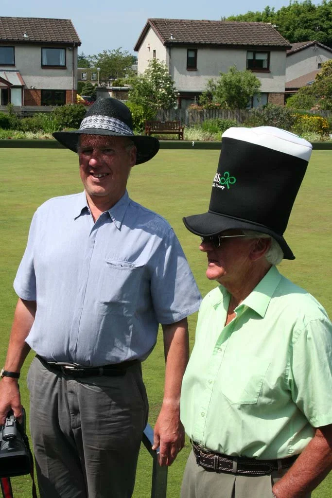 Two elderly men standing on a lawn with houses and trees in the background. One man is wearing a large Guinness beer hat and sunglasses, the other a black hat with a band and a light green shirt. The man with the Guinness hat is smiling.