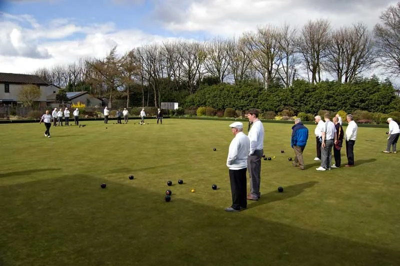 People playing lawn bowls on a green field, with trees and houses in the background under a partly cloudy sky.