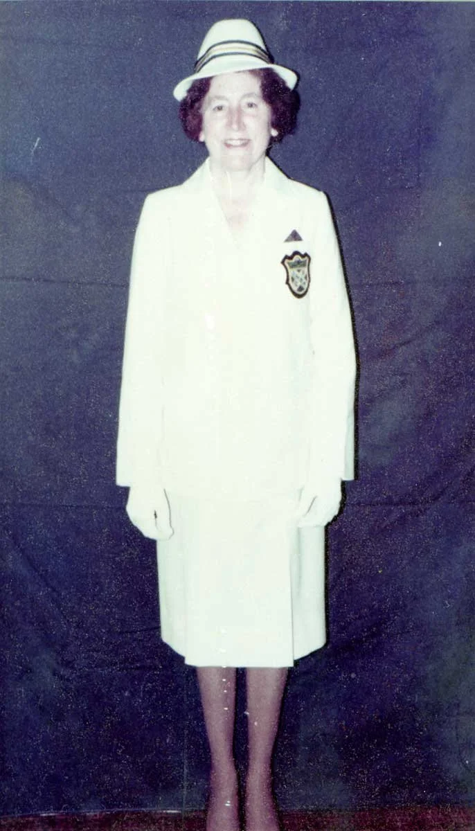 A woman smiling, dressed in a white suit and a white hat with black stripes, standing against a dark background.