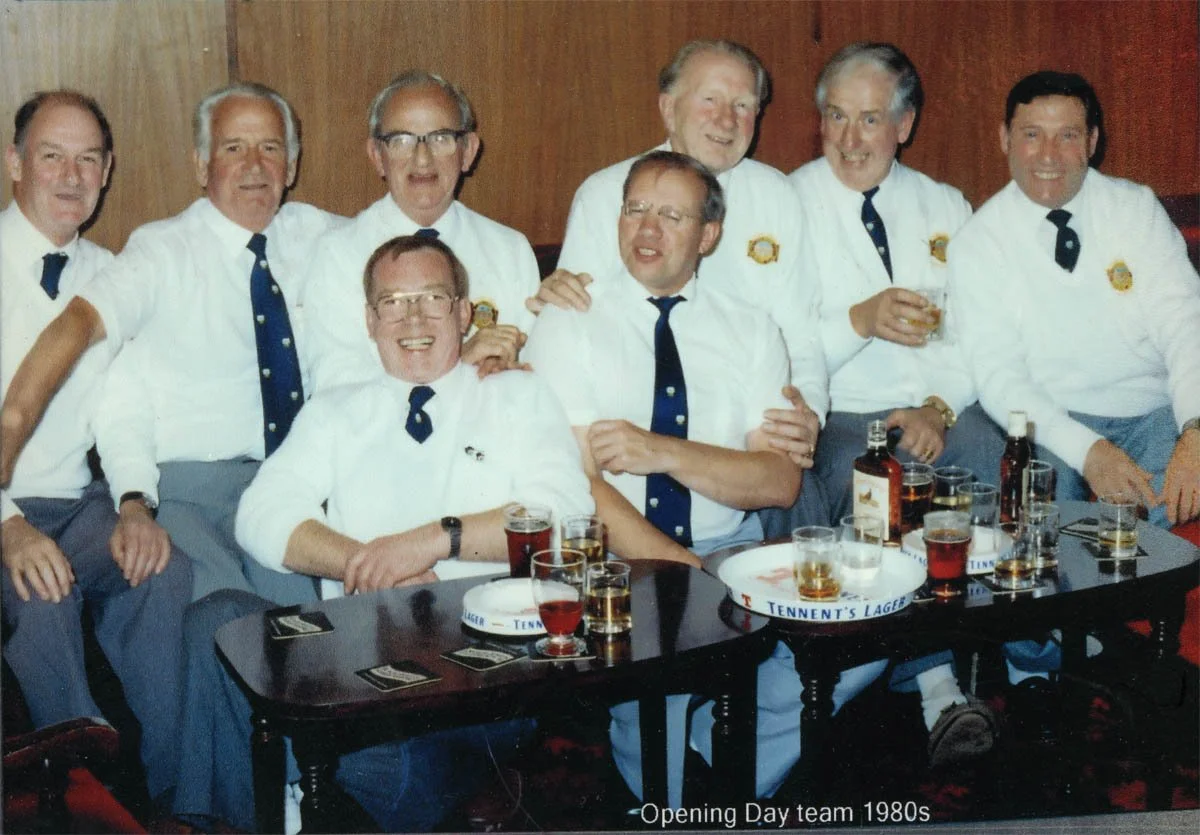 Group of nine men in white shirts, navy ties, and gray pants, sitting and standing around a table with drinks, celebrating during a gathering in the 1980s, labeled as 'Opening Day team 1980s'.