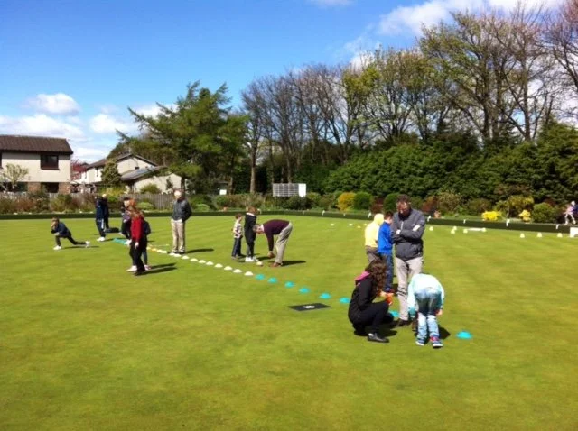 People playing lawn croquet on a green field with houses and trees in the background, sunny weather.
