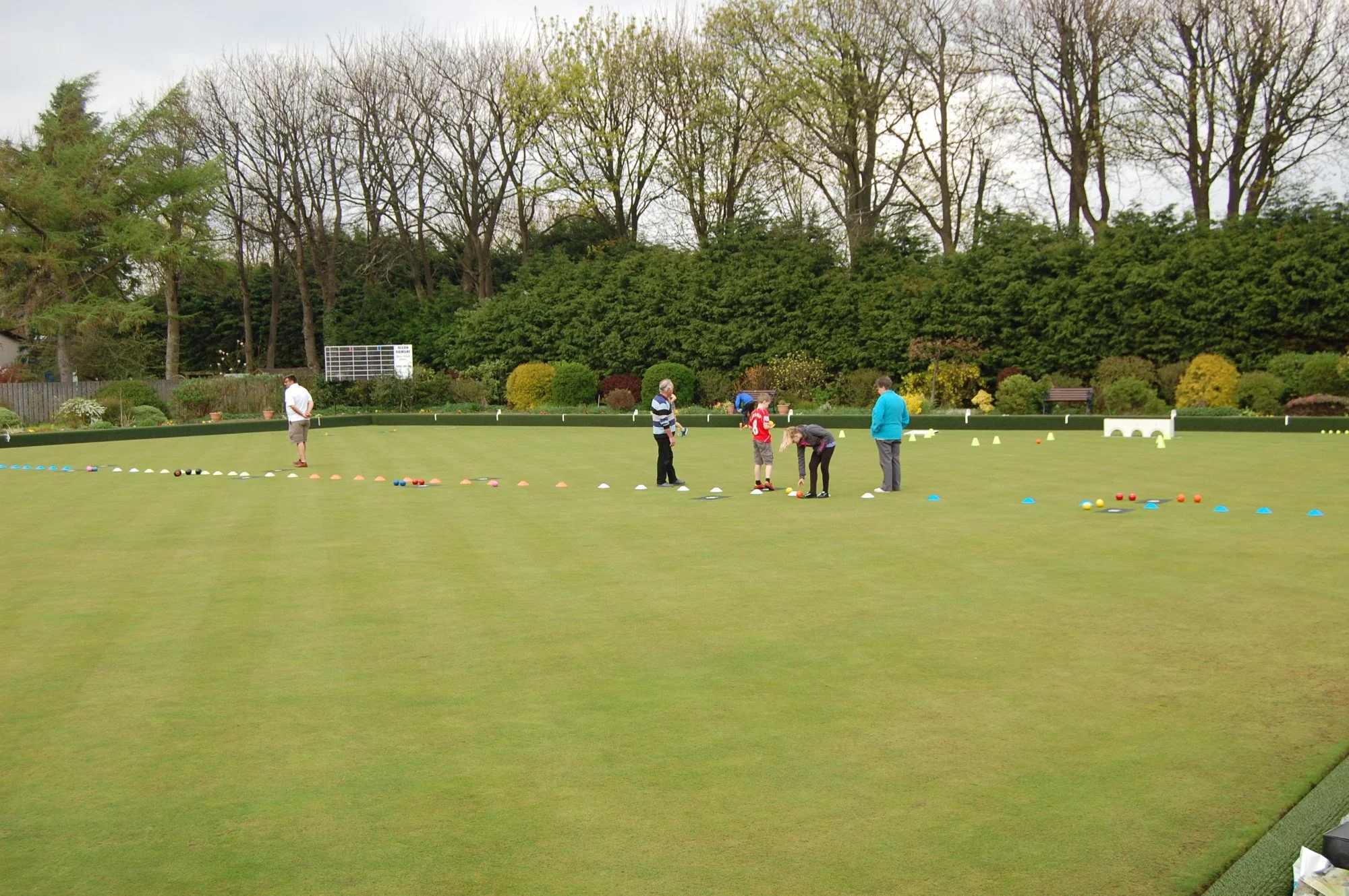 People gathered on a lawn, engaging in a game or sport involving colorful balls and cones, with trees and bushes in the background.