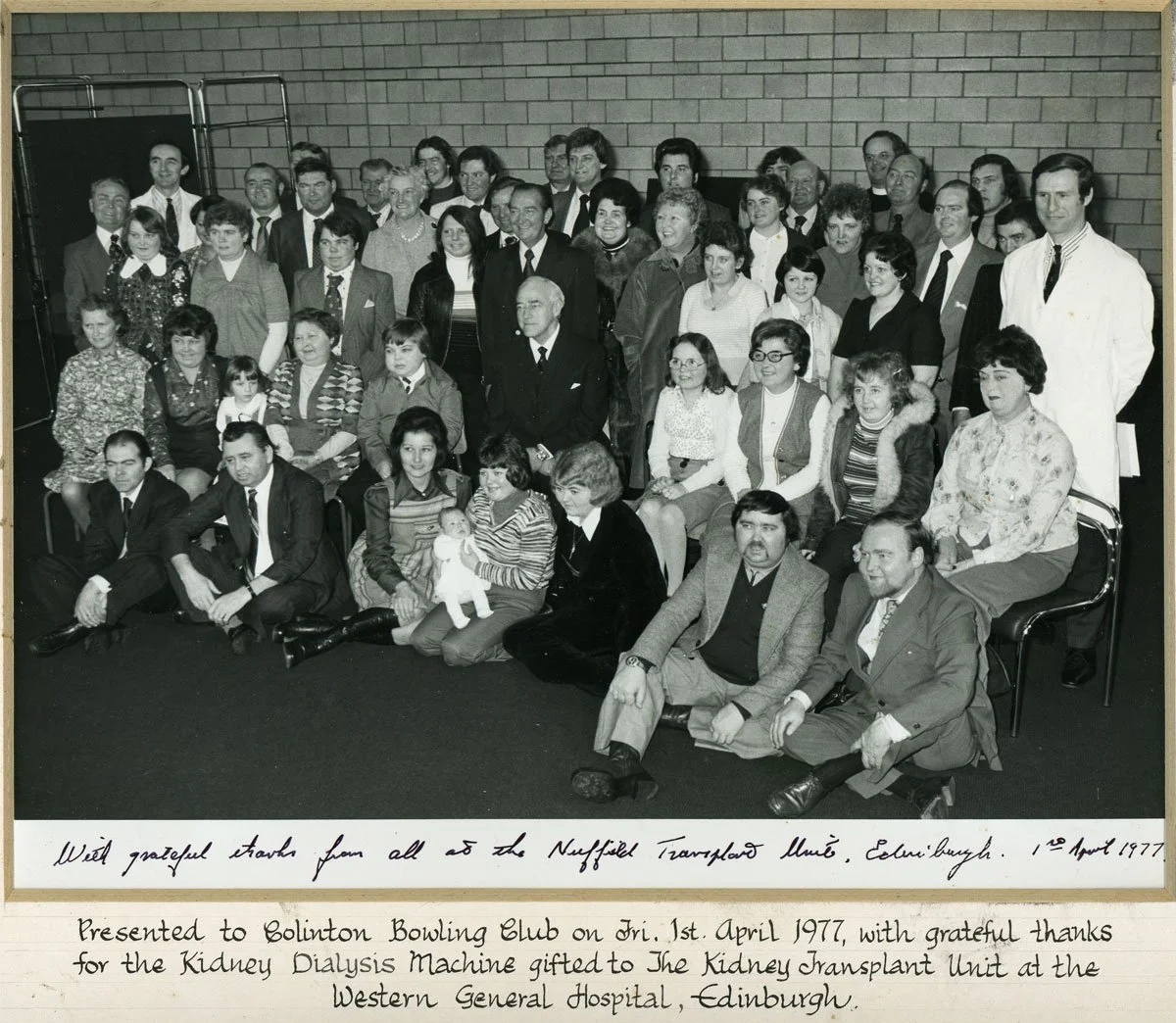 Black and white group photo of people gathered at a presentation, with handwritten notes on the bottom mentioning the presentation date and purpose related to kidney dialysis machine donation in Edinburgh in 1977.