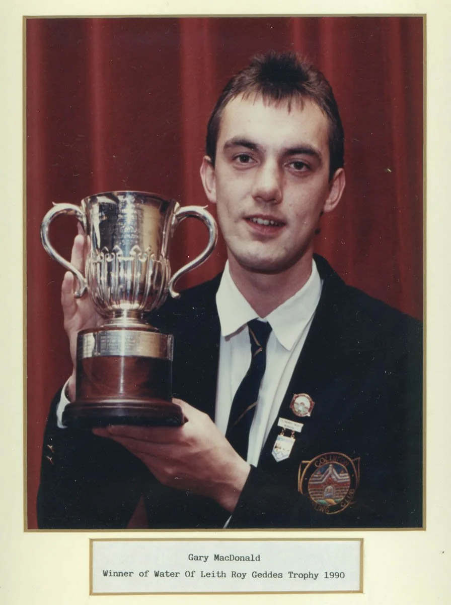 A young man in a school uniform holding a trophy, standing in front of a red curtain. The caption below reads: 'Gary MacDonald, Winner of Water Of Leith Roy Geddes Trophy 1990'.