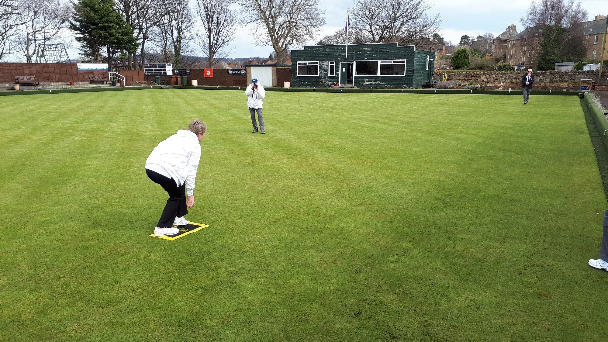 People playing lawn bowls on a well-maintained grassy bowling green, with a small green building and trees in the background.