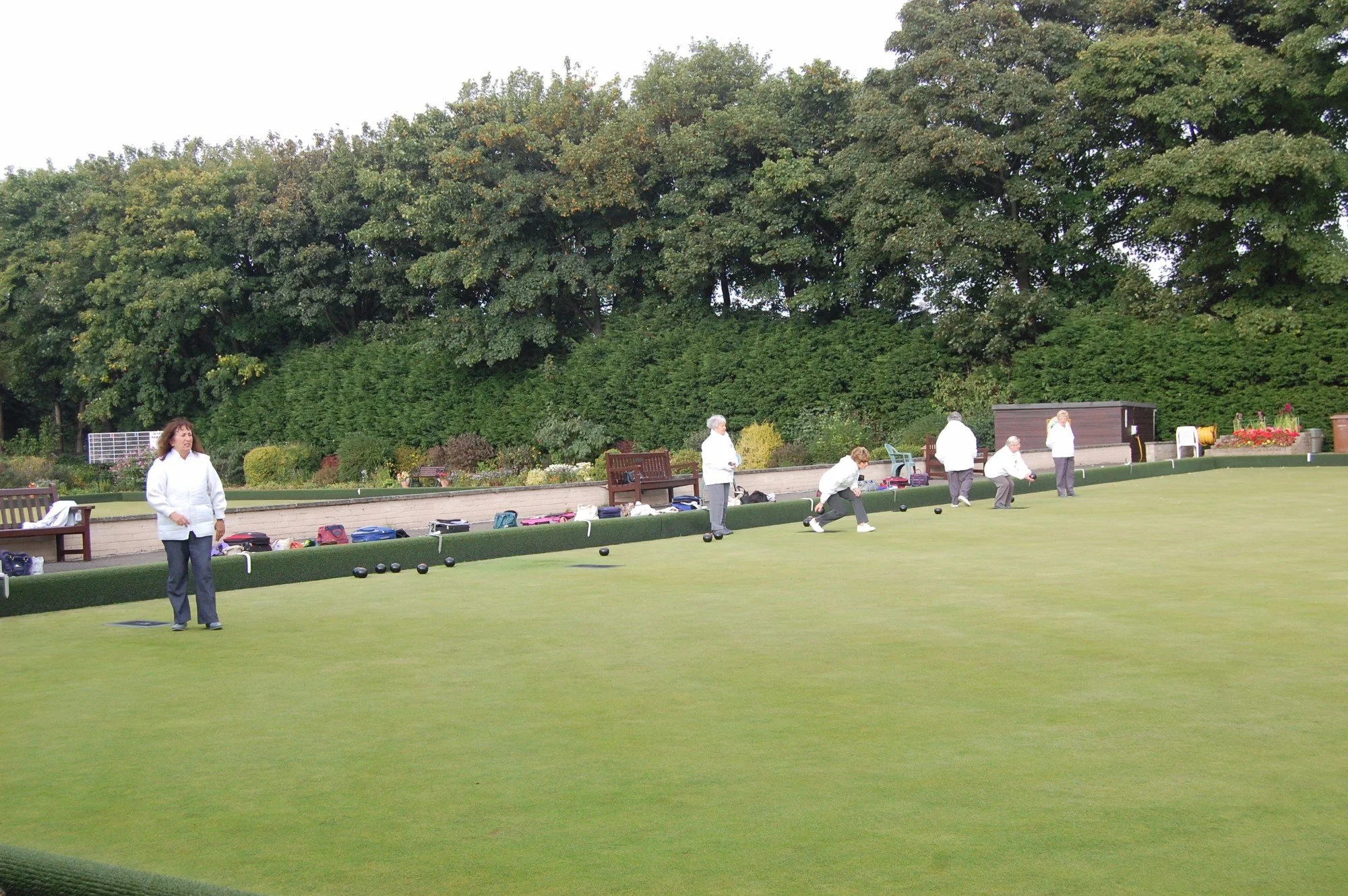 People playing lawn bowls on a green, with bags and benches along the side, and trees in the background.