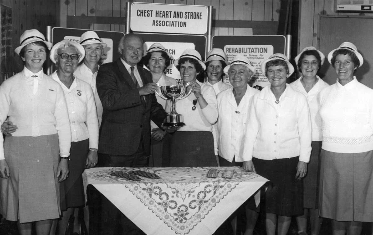 A group of women and one man celebrating with a large trophy at a Chest Heart and Stroke Association event, wearing white tops and hats, standing behind a table with a lace cloth and medals displayed.
