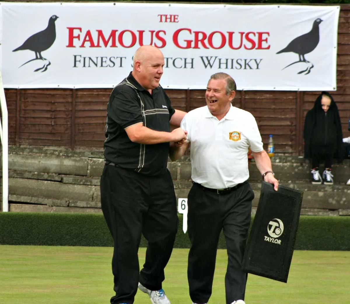 Two men smiling and shaking hands on a lawn, with a banner advertising "The Famous Ghouse" whiskey behind them, and a person in a black hoodie in the background.