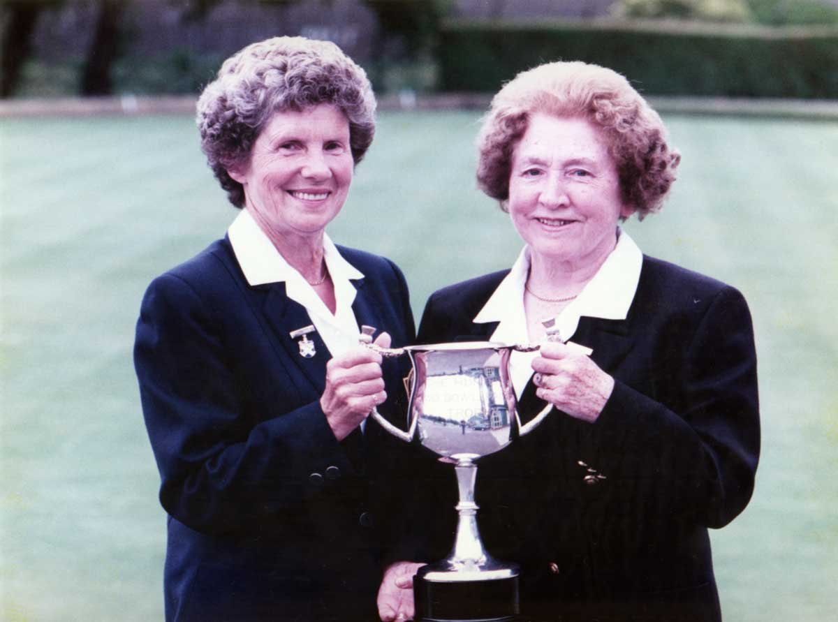 Two elderly women in navy blazers holding a silver trophy cup outdoors on a green field, smiling at the camera.