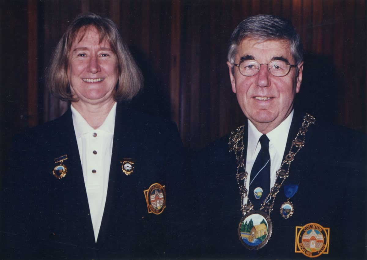 A woman and a man in formal attire, wearing badges and medals. The woman has shoulder-length light brown hair, and the man has gray hair, glasses, and is wearing a large medallion around his neck.