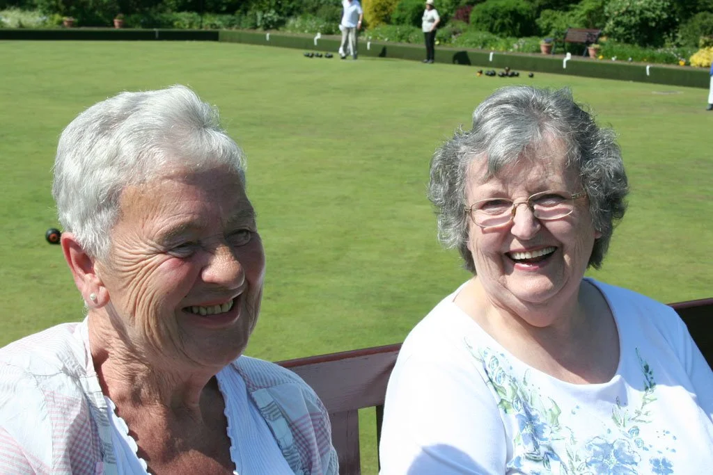 Two elderly women smiling and sitting outdoors near a lawn bowling green, with people playing in the background.