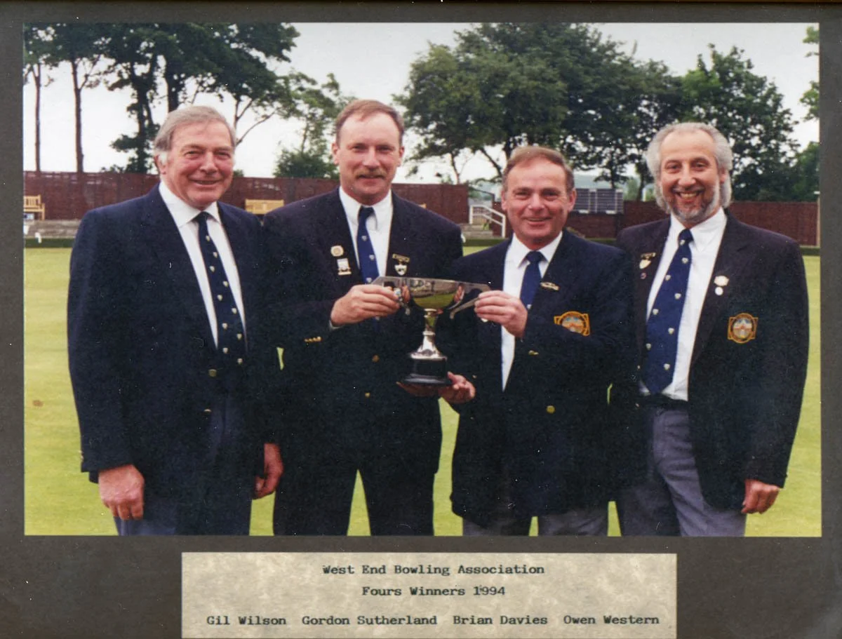 Four men in suits holding a trophy, standing on a lawn with trees in the background, celebrating their victory in the West End Bowling Association Fours in 1994.