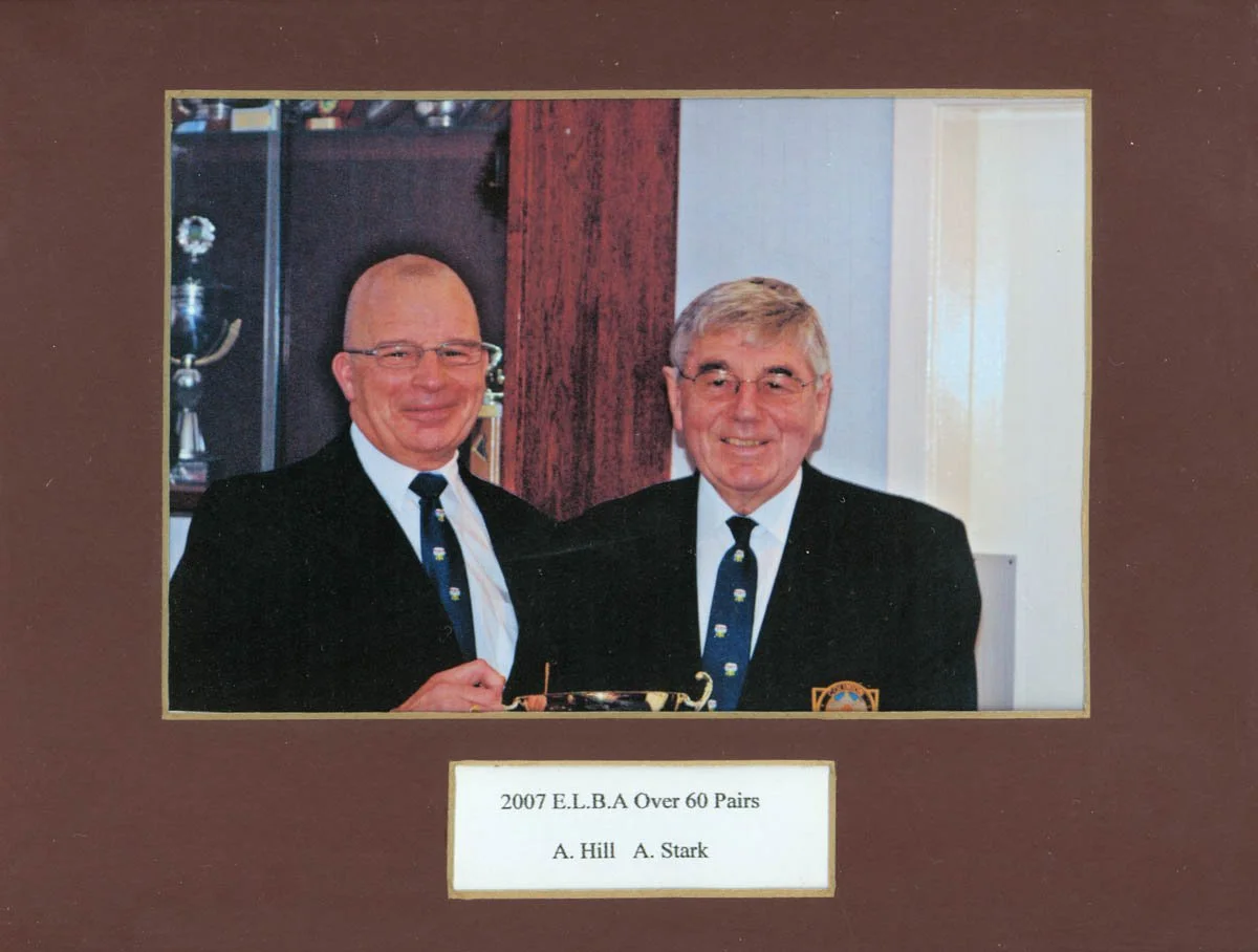 Photograph of two elderly men in formal attire with ties, smiling and standing close together at an event, with a plaque below indicating 2007 E.L.B.A Over 60 Pairs, A. Hill and A. Stark.