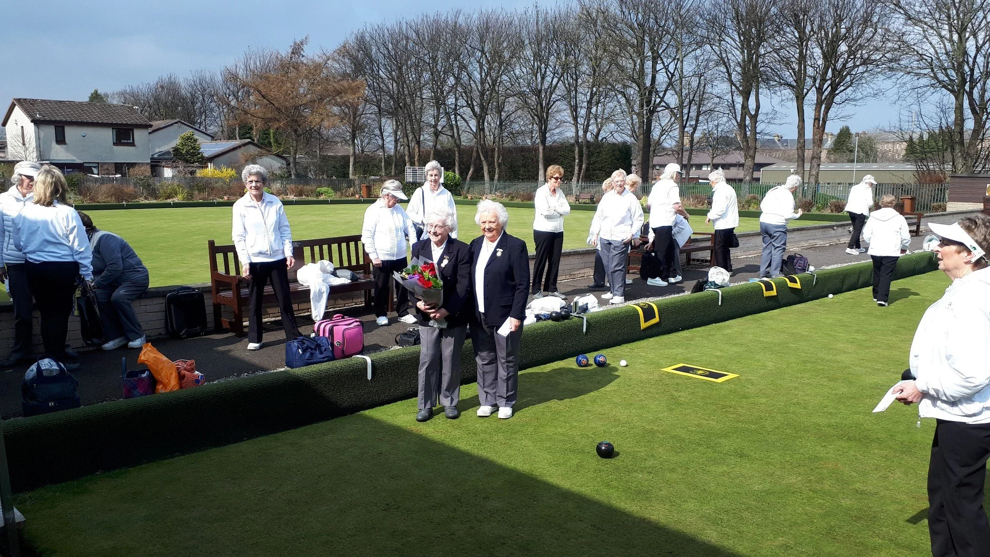A group of elderly women gathered on a lawn bowling green, with some preparing to bowl and others watching. Two women in the center are smiling; one holding a bouquet of flowers. The scene is outdoors on a sunny day with trees and houses in the backg