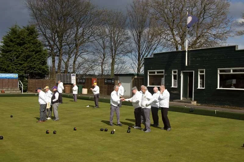 People playing lawn bowls, some exchanging awards or medals, on a well-manicured lawn with a building and trees in the background.