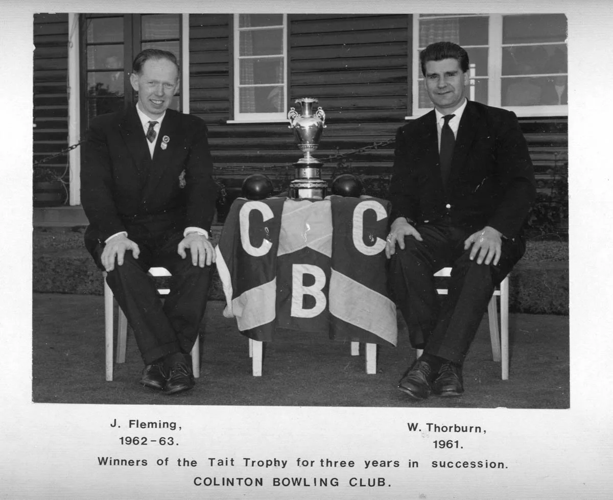 Black and white photo of two men in suits sitting on chairs with a trophy and a flag with 'C C B' hanging between them, in front of a house. The caption reads: J. Fleming, 1962-63, winners of the Tait Trophy for three years in succession. W. Thorburn