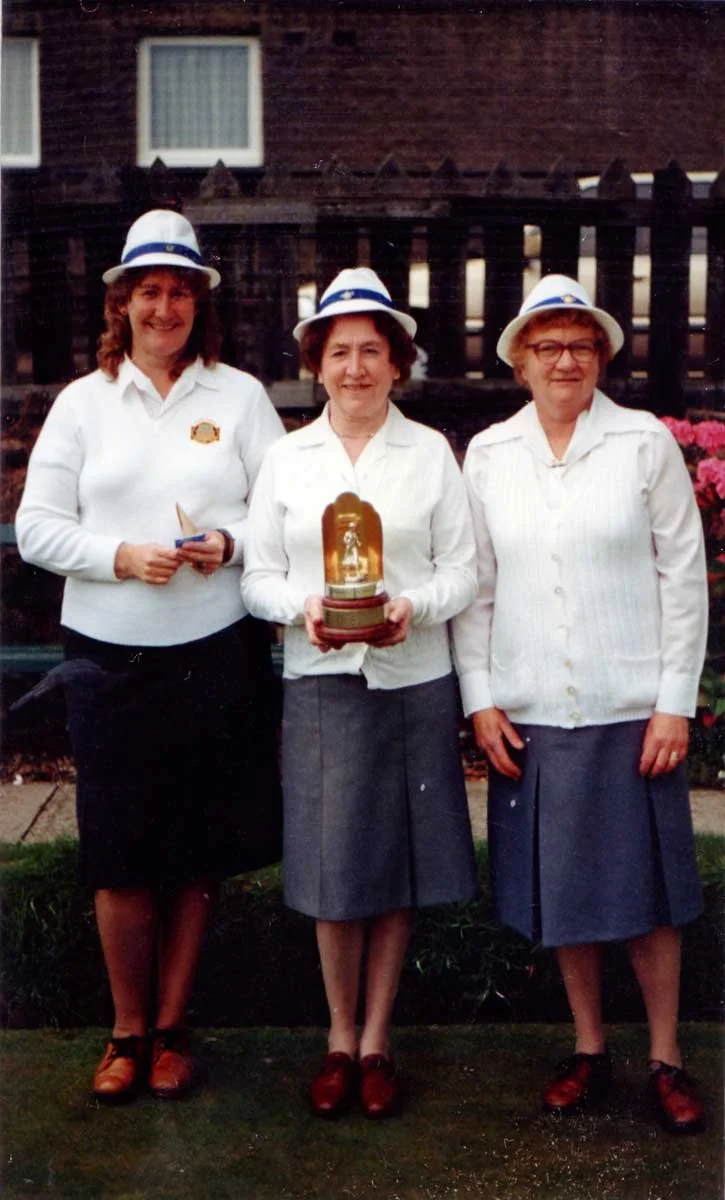 Three women dressed in white shirts, gray skirts, and white hats with blue bands, standing outside in front of a wooden fence. The woman in the center is holding a glass plaque or trophy. All three are smiling.