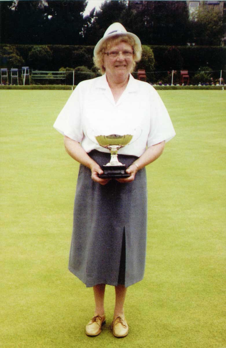 A woman standing on a green field holding a trophy, wearing a white blouse, gray skirt, beige shoes, and a white hat, with trees in the background.