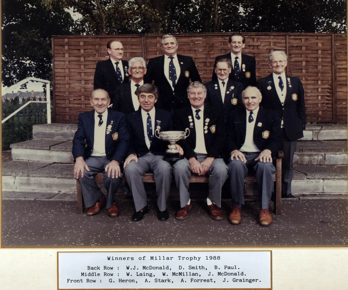 A group of eleven men in formal suits, sitting and standing on steps and a bench, celebrating with a trophy. They are members of a team that won the Millar Trophy in 1988, as indicated on the photo description.