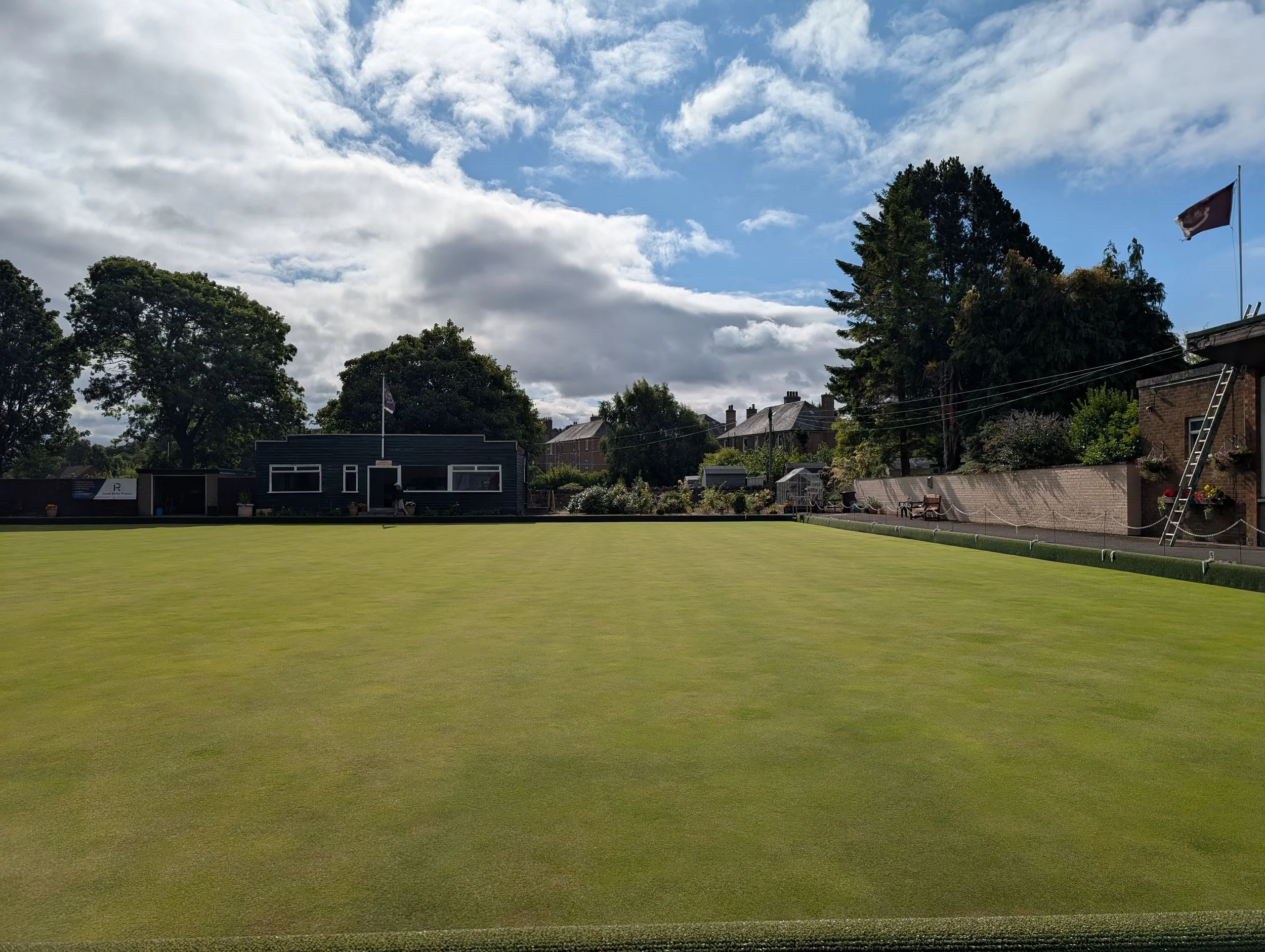 A lush green lawn with a row of trees in the background, houses, and a partly cloudy sky with patches of blue.