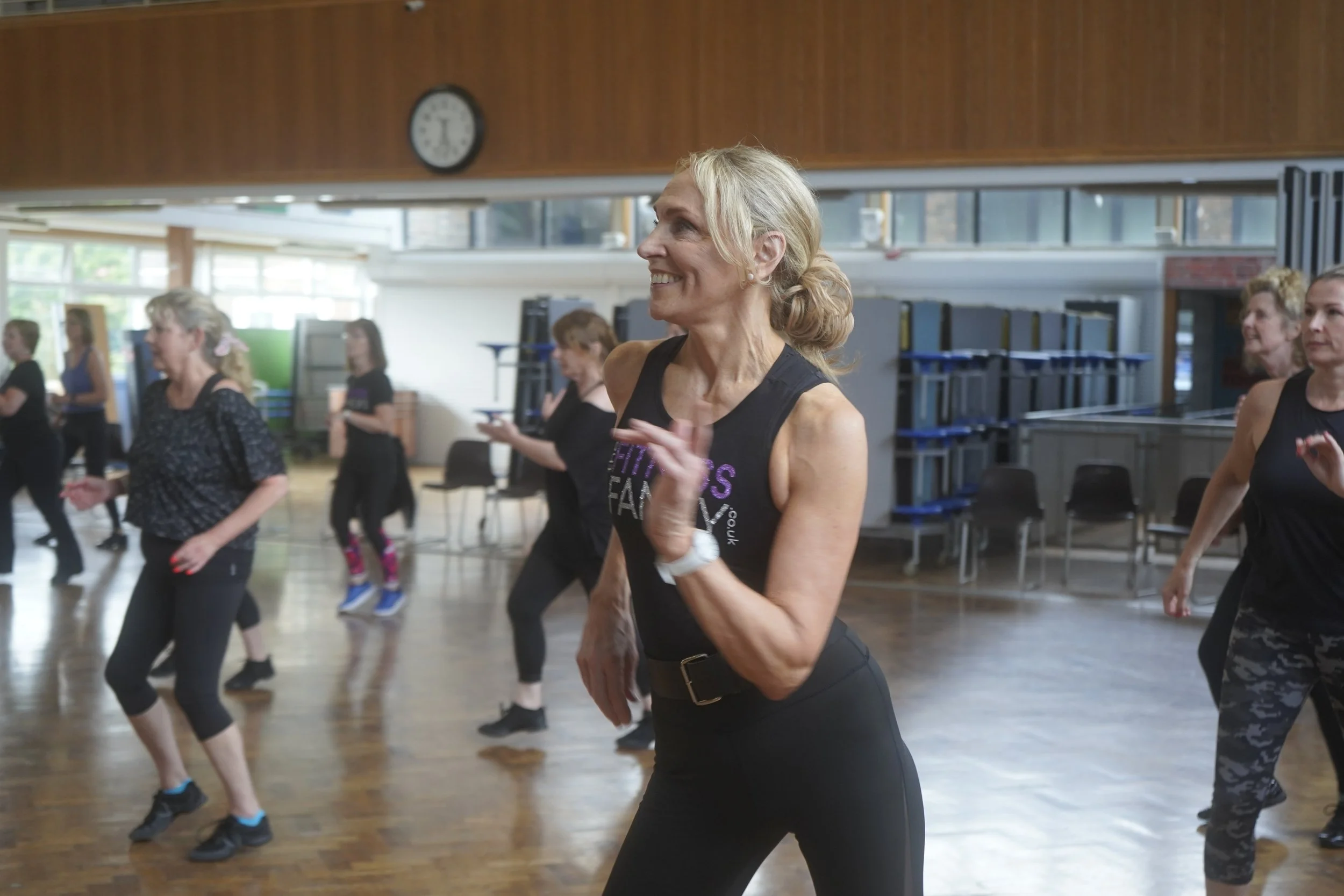A group of women participating in a fitness or dance class in a gym or community center, with a woman in the foreground smiling and moving.
