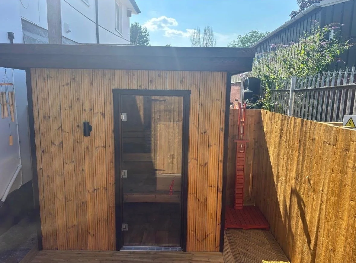 Wooden outdoor shower cabin with glass door and wooden bench inside, located on a deck next to a yellow fence and outdoor shower equipment, under a sunny sky.