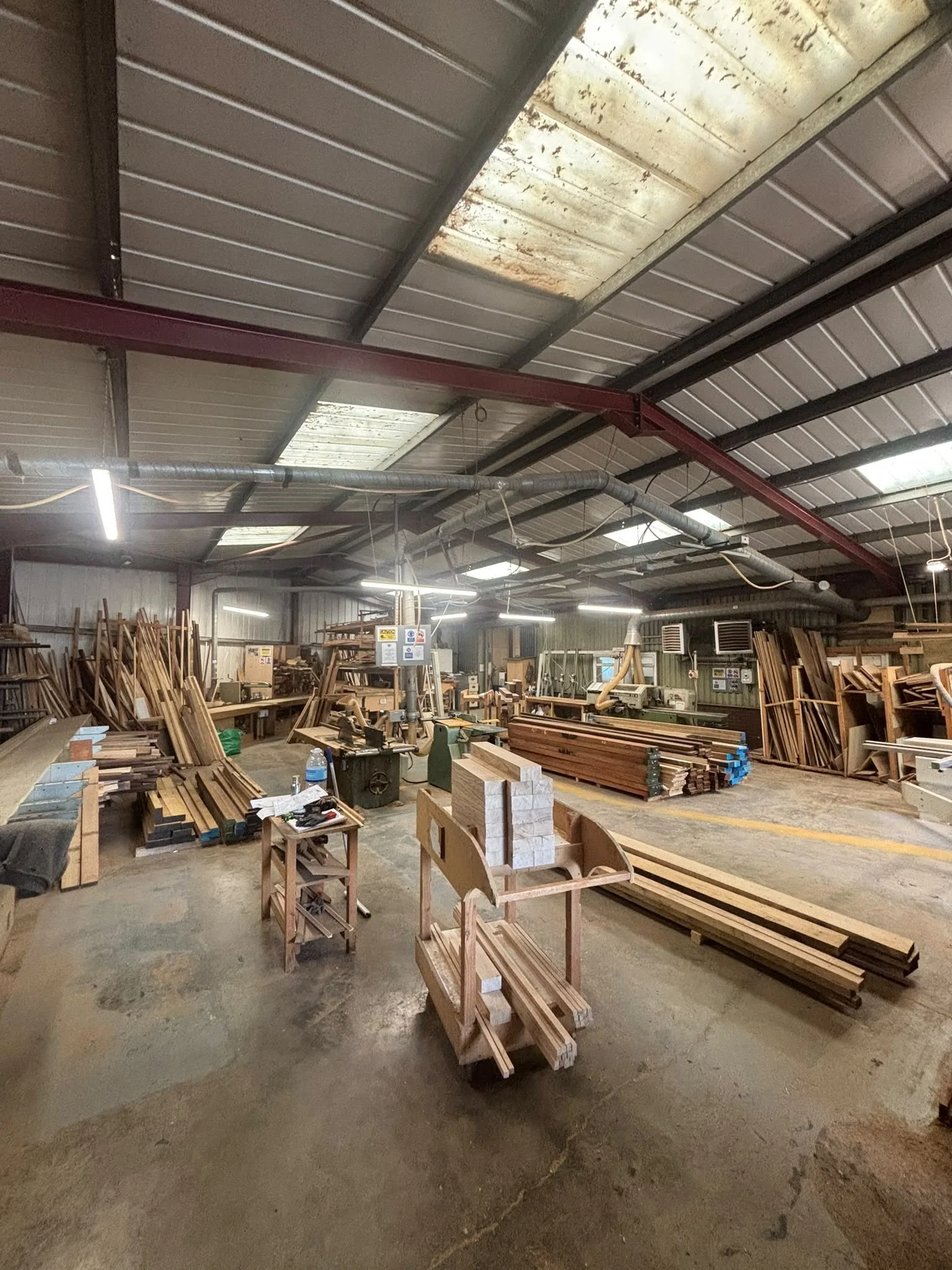 Inside a woodworking shop with stacks of lumber, workbenches, and tools, illuminated by industrial lights with a metal ceiling and ductwork.
