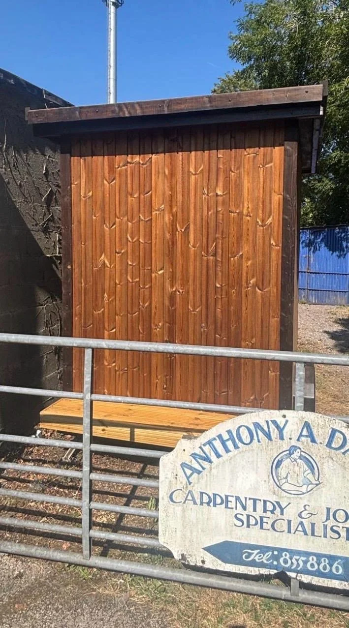 A small wooden structure with vertical wood panels, located behind a metal fence. There is a sign for Anthony A. D. Carpentry & Joinery Specialists, with a phone number, in front of the structure. A tree and blue sky are visible in the background.